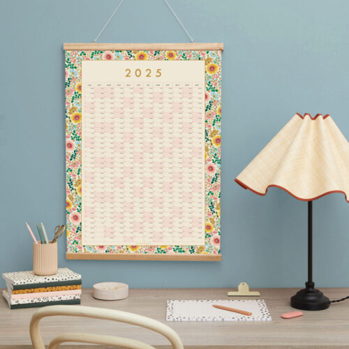 a styled desk shot of a 2025 floral wall planner with desk lamp and blue walls