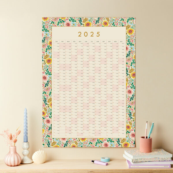a styled shot of a floral 2025 wall planner over a shelf with other matching stationery items on show on a neutral wall background