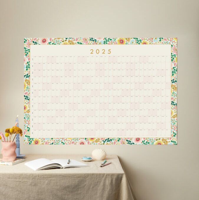 a styled shot of a large 2025 floral border year planner with table styled like a desk in front of it and neutral background wall colour