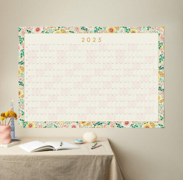 a styled shot of a large 2025 floral border year planner with table styled like a desk in front of it and neutral background wall colour