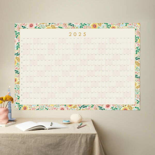 a styled shot of a large 2025 floral border year planner with table styled like a desk in front of it and neutral background wall colour