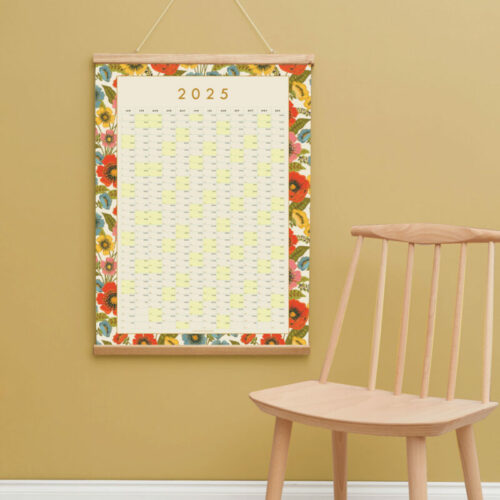 a styled shot of a 2025 brigth floral garden bloom wall planner with a light wooden chair in the front of the shot with an earthy yellow wall colour behind