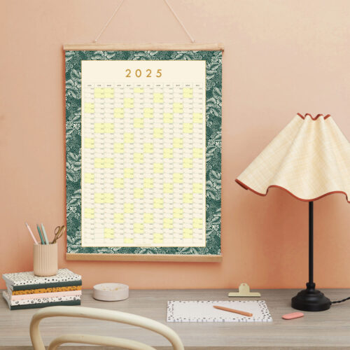 a styled desk top image of the 2025 dark green botanical wall planner the desk has pretty stationery as well as a desk lamp on it and a soft pinky background wall