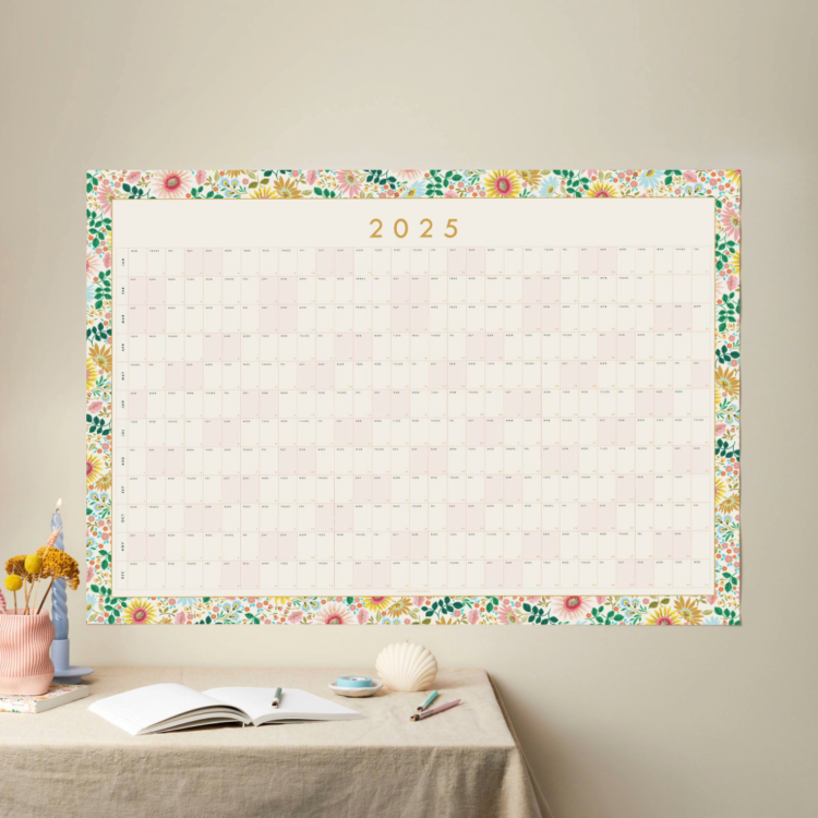 a styled shot of a large 2025 floral border year planner with table styled like a desk in front of it and neutral background wall colour